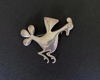 Puede incluir: Un broche de metal plateado con forma de pájaro estilizado en movimiento. El pájaro presenta una cabeza redondeada con un ojo circular, un ala triangular con un pequeño detalle grabado, una cola de tres lóbulos y dos patas dobladas al correr.