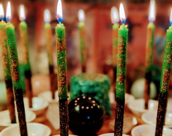 May include: A circle of green and black candles on a wooden surface with small colourful stones. The candles are in white ceramic dishes.