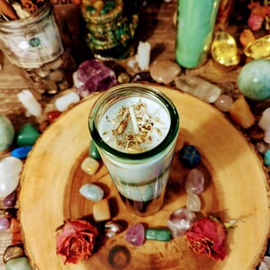 May include: A white candle with gold flakes embedded in the wax sits on a wooden tray surrounded by colorful stones. The candle is in a clear glass jar with a green base.