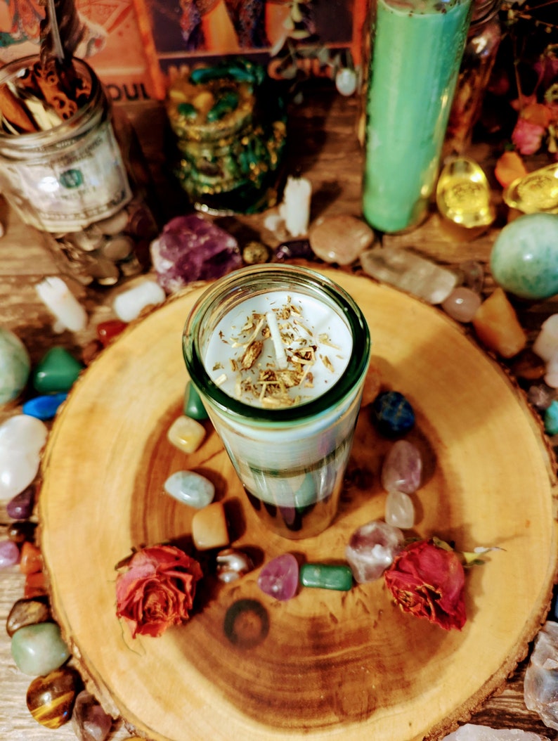 May include: A white candle with gold flakes embedded in the wax sits on a wooden tray surrounded by colorful stones. The candle is in a clear glass jar with a green base.