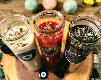 May include: Three glass jar candles with eye designs, each with a different colour wax: white, red, and black. The candles are on a wooden slice surrounded by colourful stones. The candles are decorated with herbs and other natural elements.