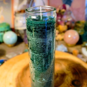 Puede incluir: Una vela de vidrio verde en un frasco de vidrio transparente con una mecha blanca. La vela está sobre una superficie de madera.