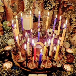 Puede incluir: Una bandeja circular de madera contiene numerosas velas cónicas encendidas de varios colores, incluyendo morado, amarillo, negro y rojo, colocadas en pequeños recipientes de color terracota. Las velas tienen texturas moteadas y follaje verde.