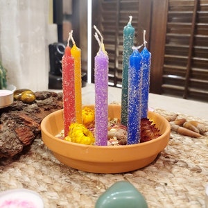 May include: A set of seven small candles in a rainbow of colors, arranged in a terracotta pot with dried flowers. The candles are surrounded by small stones and dried flowers.