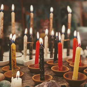 May include: A collection of lit candles in various colors, including red, black, yellow, and white, are arranged in small terracotta dishes on a wooden surface. The candles are surrounded by small stones and crystals.
