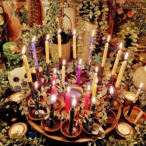 May include: A collection of lit candles in various colors, including purple, pink, and yellow, arranged on a wooden tray with small terracotta holders. The candles are surrounded by greenery, creating a mystical and decorative display.
