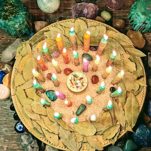 May include: A gold tray with a circle of bay leaves surrounding a collection of colorful candles, crystals, and a small white candle in the center. The tray is surrounded by more crystals and a wooden surface.