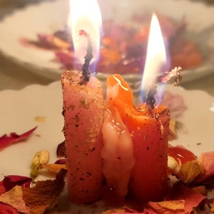 May include: Three lit candles with orange and pink wax, surrounded by dried rose petals and other botanicals. The candles are on a white plate with a floral design. The flames are bright and the scene is softly lit.