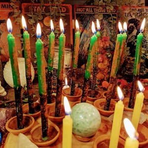 May include: A collection of green and yellow candles burning in small terracotta pots. The candles are arranged in a circle around a green crystal ball. The candles are surrounded by tarot cards with the text "Stay Wild", "Not All Those Who Wander Are Lost", and "We Are The Daughters Of The Moon".