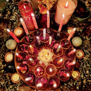 Op de afbeelding: Een rond houten dienblad bevat talloze kleine kaarsenhouders met brandende taps toelopende kaarsen, verspreide gedroogde rozenblaadjes en een centrale votiefkaars. Een houten kalender toont de datum 07 augustus 2026 en www.enhergyintentions.com.