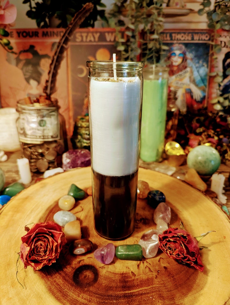 May include: A white and black candle in a glass jar with a gold lid. The candle is surrounded by colorful stones and two dried roses on a wooden surface.