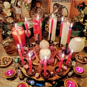 May include: A spiritual display with lit red candles in holders, encircled by crystals, dried flowers, and cinnamon sticks on a wooden base. Several glass jar candles and a white spherical candle create a warm, inviting atmosphere. The scene evokes a sense of ritual.