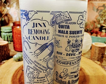 May include: Two tall white wax candles in clear glass cylinders featuring blue printed graphics and text. The left candle is labelled JINX REMOVING CANDLE with illustrations of a bat, snake, magnet, dice, and cat. The right candle is labelled QUITA MALA SUERTE.