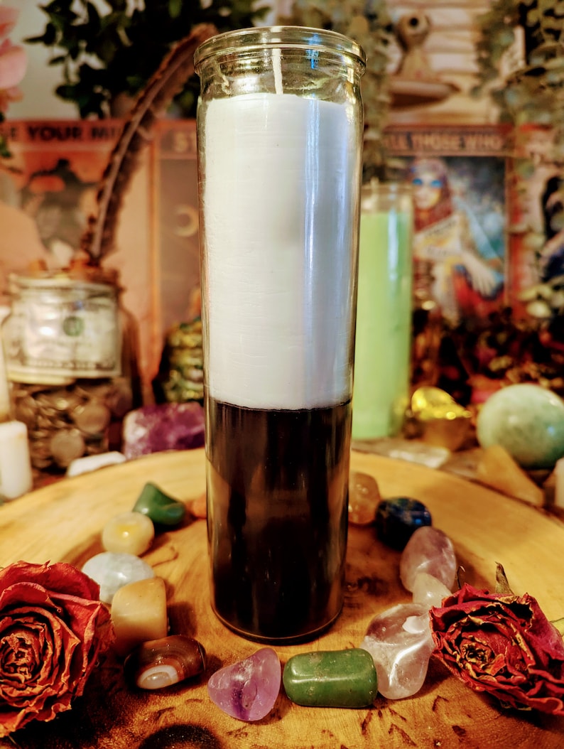 May include: A white and black candle in a clear glass jar. The candle is surrounded by colorful stones and dried roses.