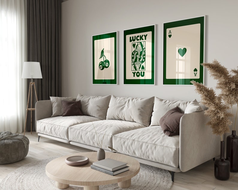 Puede incluir: Un sof&aacute; blanco con tres almohadas se encuentra frente a una pared con tres impresiones enmarcadas. Las impresiones muestran una cereza verde, una carta de juego verde con el texto "Lucky You" y un as de picas verde. Una planta en maceta con hojas altas y plumosas se encuentra en el suelo a la derecha del sof&aacute;.