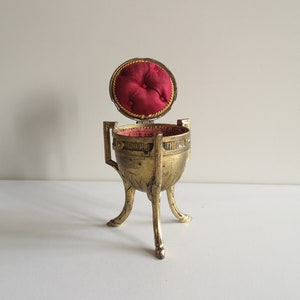 May include: A gold-colored trinket box with a red velvet interior. The box is shaped like an egg and sits on three legs.