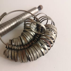 Op de afbeelding: Een set zilverkleurige metalen ringmaten met op elke ring gegraveerde nummers. Een metalen meetlat is verbonden met de ringmaten. De ringmaten worden gebruikt om vinger maten voor ringen te meten.