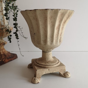 Könnte beinhalten: Ein kleiner, weißer, gusseiserner Pflanzer mit geriffeltem Design und einem quadratischen Sockel mit vier Füßen. Der Pflanzer ist im Vintage-Stil gehalten und hat eine abgenutzte Oberfläche.