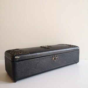 May include: A long, rectangular black box with silver accents and a decorative lock. The box has a textured surface and appears to be made of leather.