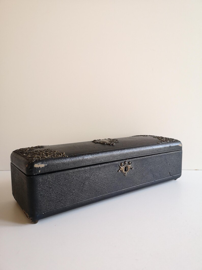 May include: A long, rectangular black box with silver accents and a decorative lock. The box has a textured surface and appears to be made of leather.