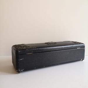 May include: A black rectangular box with a decorative metal design on the top and a hinged lid. The box is made of a textured material and has a worn look.