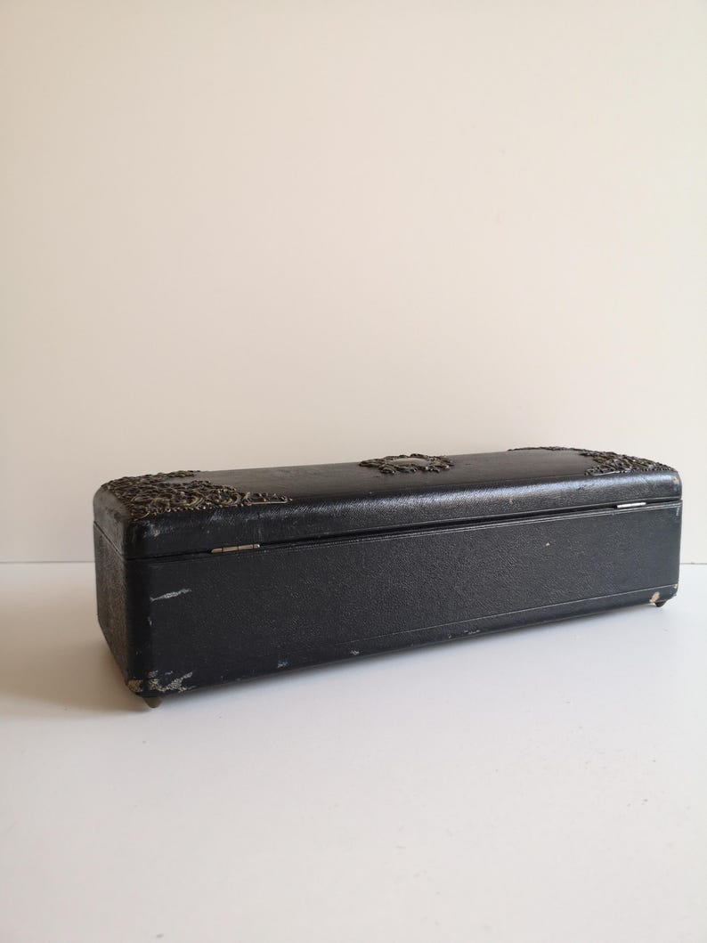 May include: A black rectangular box with a decorative metal design on the top and a hinged lid. The box is made of a textured material and has a worn look.