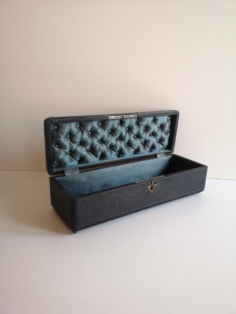 May include: A black rectangular box with a tufted blue velvet lining. The box has a metal clasp and is lined with a blue fabric.