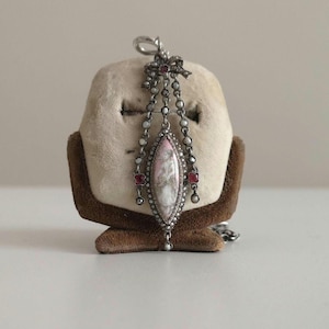 May include: A silver pendant with a pink and white cameo, surrounded by small pearls and red gemstones. The pendant hangs from a chain with a bow at the top.