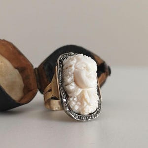 Può includere: Un anello d'oro con un cammeo rettangolare color avorio raffigurante un volto scolpito. Il cammeo è incorniciato da un metallo color argento con piccole pietre trasparenti. L'anello è posto su uno sfondo neutro.