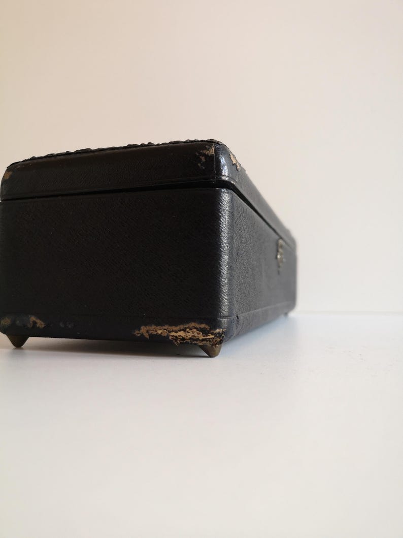 May include: A black rectangular box with a worn leather exterior and gold metal feet. The box is closed and has a slight angle.