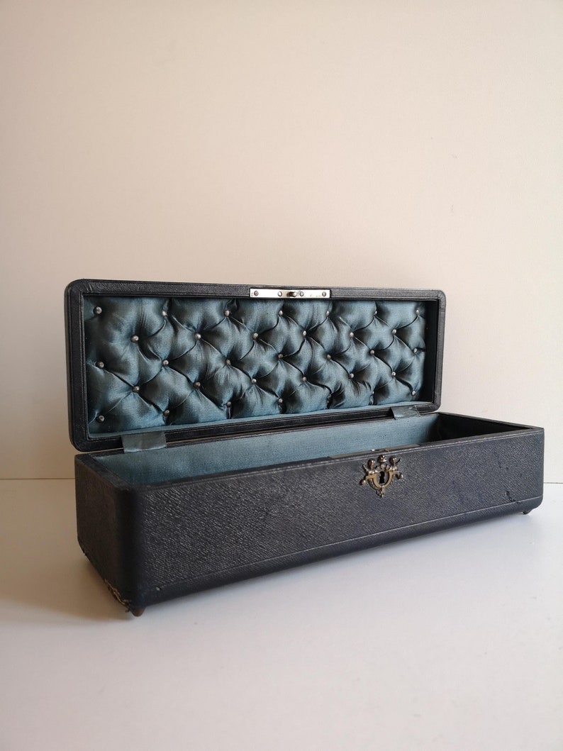 May include: A black rectangular box with a tufted blue fabric lining and a metal latch. The box is open and the interior is visible.