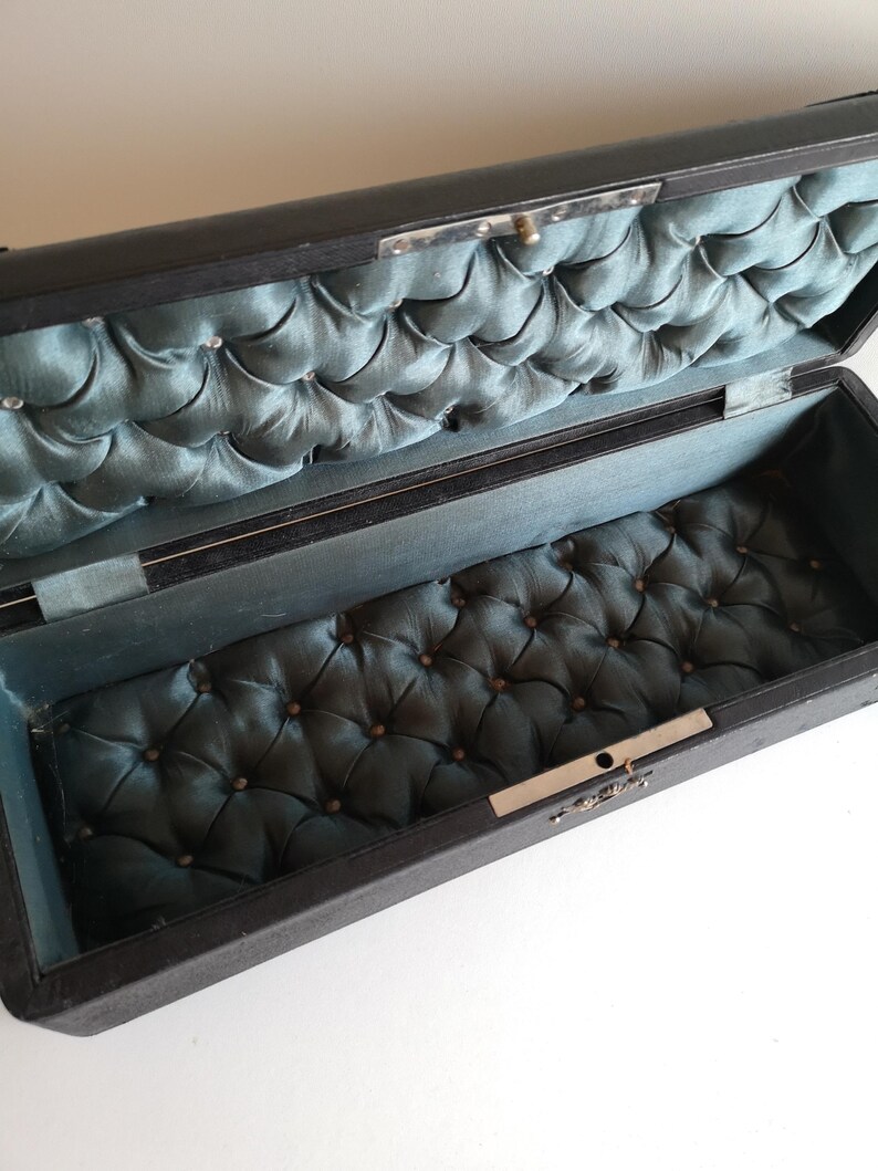 May include: A black rectangular box with a hinged lid. The inside of the box is lined with a blue quilted fabric.
