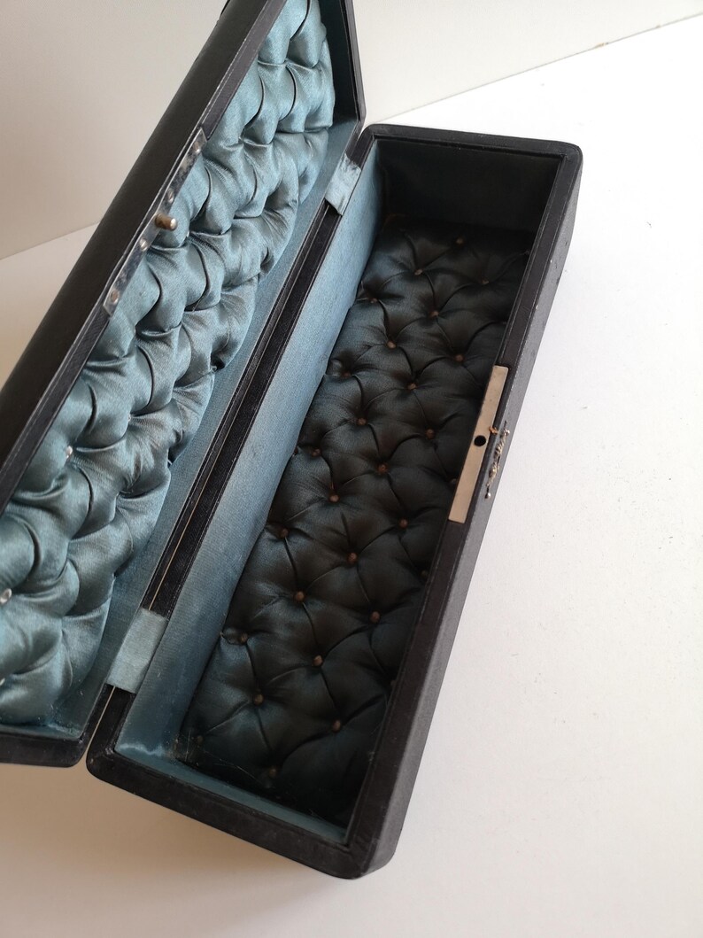 May include: A black rectangular box with a hinged lid. The inside of the box is lined with a dark blue fabric with a tufted pattern.
