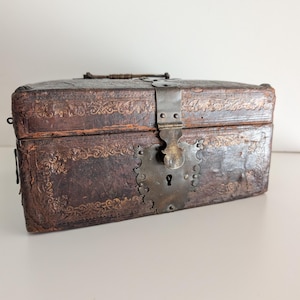 May include: A rectangular dark brown leather box featuring an embossed gold-toned floral border pattern. The box includes a metal handle on the top and a large, ornate, heart-shaped metal latch mechanism with a central keyhole on the front.