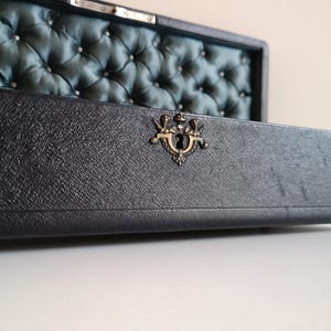 May include: A vintage wooden box with a dark blue leather exterior and a teal blue satin interior. The box has a brass latch with a keyhole and a tufted design on the inside.