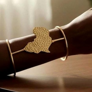 May include: Gold-toned bracelet featuring a textured map-shaped charm. The bracelet has a minimalist design with a thin band that wraps around the wrist. The charm is attached to the band with delicate lines.