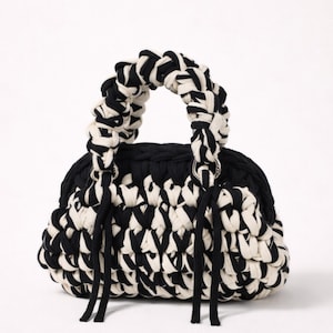 May include: A black and white woven handbag with a rounded shape. The bag features a braided handle in black and white, and black tassels hang from the base. The bag's body is a mix of black and white woven material.