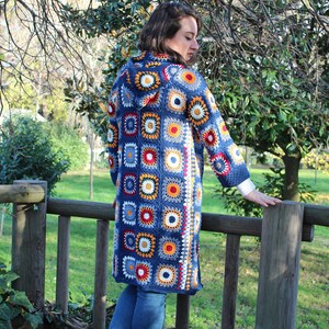 May include: A long blue crochet cardigan with a hood, featuring a colorful geometric pattern of squares and circles. The cardigan is worn over blue jeans.