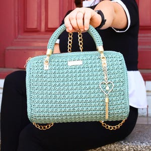 Pode incluir: Uma bolsa de crochê verde menta com alças e detalhes de corrente dourada. A bolsa retangular apresenta uma textura tecida, uma alça superior e um pingente em forma de coração com uma borla. A bolsa é segurada por uma pessoa.