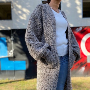 May include: A long, gray cardigan with a textured knit pattern. The cardigan has a V-neck and pockets on the front. The cardigan is worn over a white t-shirt and blue jeans.