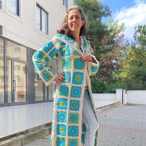 Könnte beinhalten: Ein langer, gehäkelter Cardigan mit Kapuze. Der Cardigan ist aus beigefarbenem Garn mit blauen und grünen Quadraten gefertigt. Der Cardigan wird über einer blauen Jeans getragen.