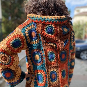 Puede incluir: Una larga chaqueta de punto de ganchillo con capucha. La chaqueta está hecha de cuadrados coloridos con fondo marrón. Los cuadrados presentan una variedad de colores, incluyendo azul, verde, naranja y rojo.