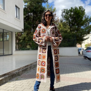 Puede incluir: Un cárdigan largo de crochet beige con un patrón geométrico colorido. El cárdigan tiene capucha y mangas largas. La persona que lleva el cárdigan está de pie en una acera.