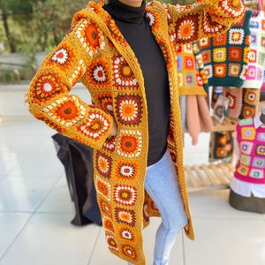 Könnte beinhalten: Ein langer, gehäkelter Cardigan mit Kapuze. Der Cardigan ist aus gelbem Garn mit orangefarbenen, braunen und weißen Akzenten gefertigt. Der Cardigan ist mit großen, bunten Quadraten gemustert.