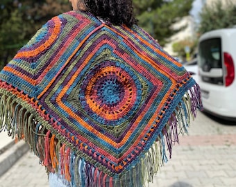 May include: A colorful, handmade crochet shawl with a square design and fringe. The shawl features concentric squares in shades of orange, blue, green, purple, and pink. The fringe is a mix of colors. The shawl is worn over the shoulders.