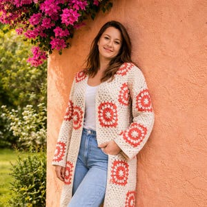 May include: Cream-colored, long crochet cardigan with red floral patterns. The open-front sweater features a relaxed fit and long sleeves. The model is wearing a white top, blue jeans, and white sneakers. The background includes greenery and a peach-colored wall.