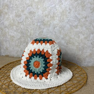 Puede incluir: Sombrero de sol de crochet blanco con un patr&oacute;n geom&eacute;trico colorido en tonos naranja, verde y azul. El sombrero est&aacute; sobre una estera de paja tejida.