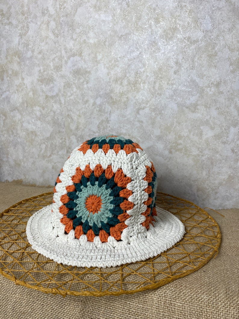 Puede incluir: Sombrero de sol de crochet blanco con un patr&oacute;n geom&eacute;trico colorido en tonos naranja, verde y azul. El sombrero est&aacute; sobre una estera de paja tejida.