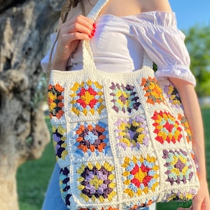 May include: A white crocheted tote bag with colorful granny squares. The bag has a long strap and is made with a variety of colors, including red, yellow, blue, green, and purple.