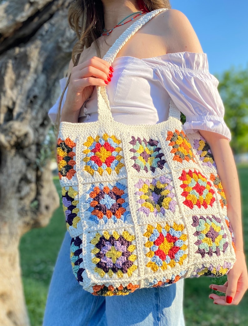 May include: A white crocheted tote bag with colorful granny squares. The bag has a long strap and is made with a variety of colors, including red, yellow, blue, green, and purple.